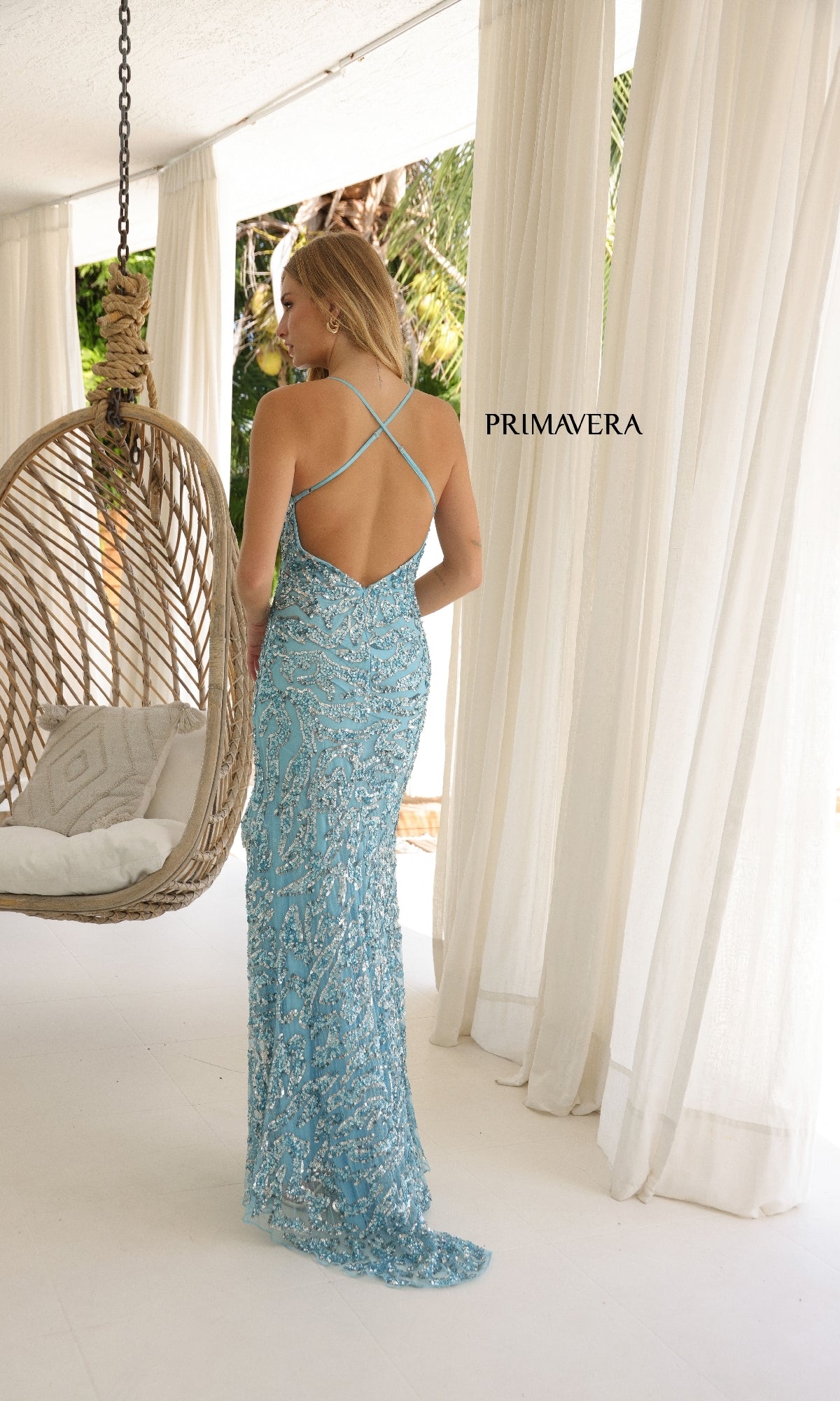 A woman stands with her back to the camera on a white patio, wearing the Primavera 4348 long prom dress with hot stone embellishments and crisscross straps, near a hanging chair and flowing curtains.